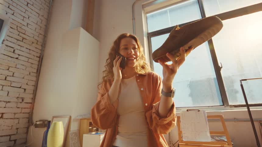 A cheerful female entrepreneur in her sunlit workshop holds up a suede shoe while confirming details over a phone call