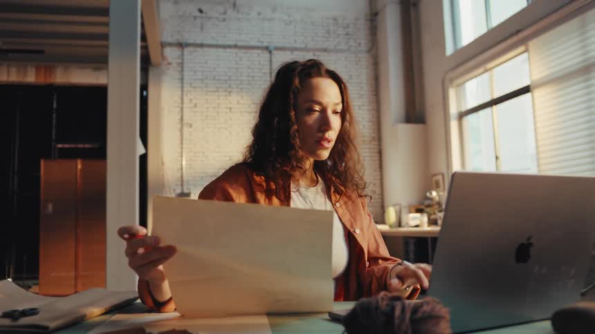 A focused female designer references her sketches while working on a laptop in her sun-drenched workshop