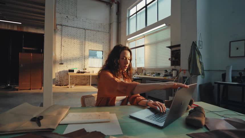 A young female artisan closes her laptop after finishing work for the day in her sunlit industrial studio