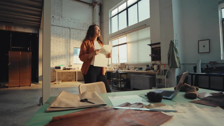 A pensive female artisan examines a paper pattern, seeking inspiration for her next creation in a sunlit industrial workshop