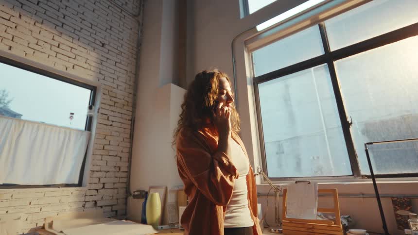 A happy young woman with curly hair talks on her phone while standing in a sunlit industrial loft studio