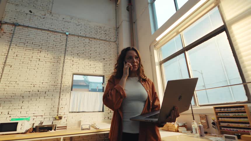 A woman multitasks by talking on her phone while holding and typing on a laptop in a sunlit industrial workshop