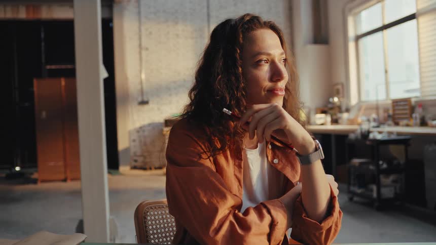 Inspired female artist daydreams peacefully in her sun-drenched creative studio during golden hour