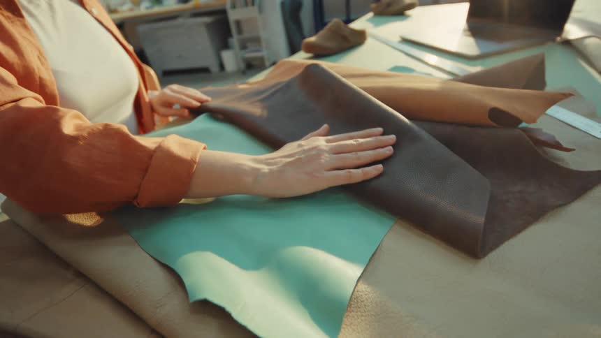 A female artisan carefully examines and touches various colorful pieces of genuine leather in her sunlit workshop