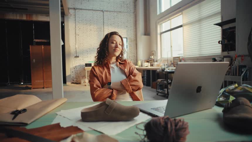 A young female shoe designer finishes her work on a laptop and relaxes, feeling a sense of accomplishment in her sunlit studio