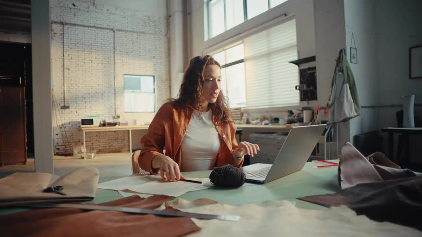 A female artisan works on a laptop and takes notes at her desk in a spacious, creative studio
