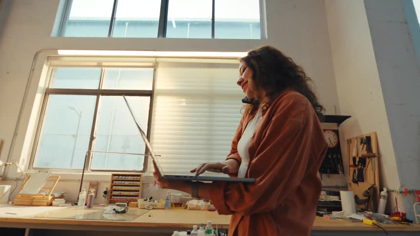 A young female artist multitasks in her sunlit studio, talking on the phone while working on her laptop
