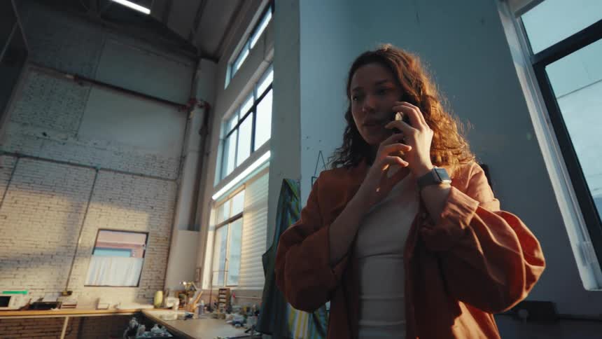 A low angle slow motion shot of a thoughtful young woman talking on her smartphone inside a sunlit industrial loft studio