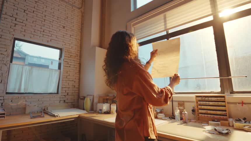 A female artist holds a sheet of paper up to the golden sunlight streaming through a large window in her creative studio