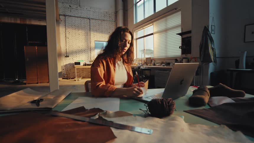 A young female fashion designer sits at a large table in her sunlit studio, working on new designs with a laptop, sketches, and leather materials