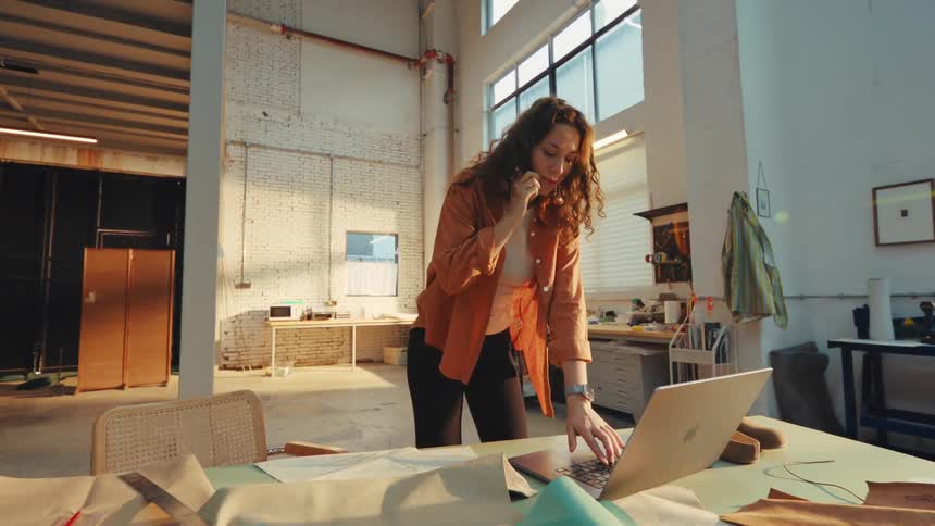 A young female artisan manages her business, talking on the phone and using a laptop in her sunlit industrial workshop