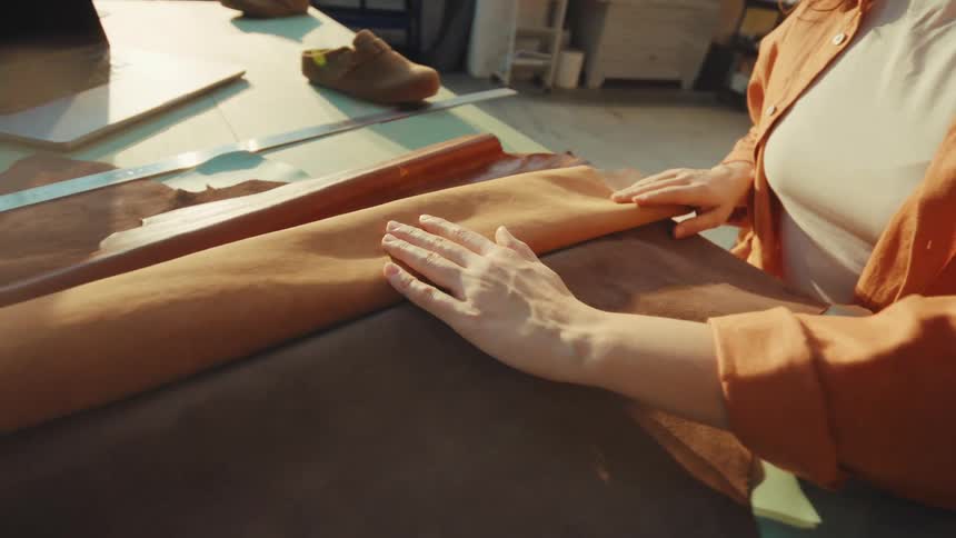 A female artisan gently unrolls and feels the texture of a piece of brown leather in her sunlit workshop