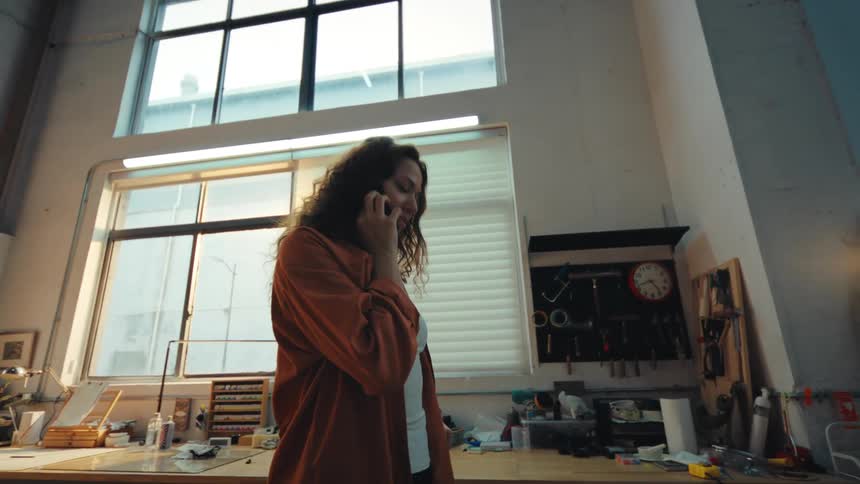 A young female artist has a dynamic phone conversation in her sunlit, industrial loft style art studio