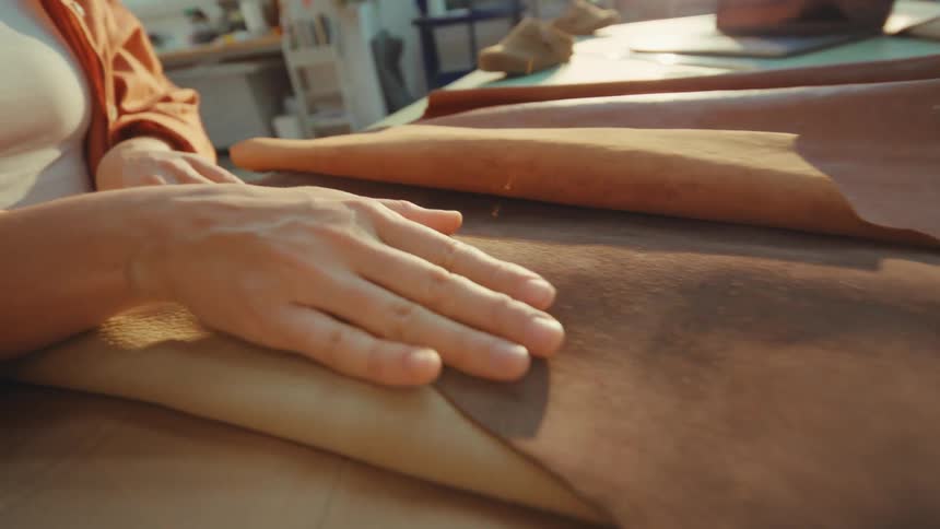 Close-up of a female artisan's hands feeling the texture of soft brown leather in a sunlit workshop