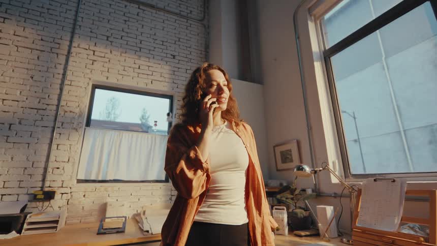 A happy woman smiles while enjoying a pleasant phone conversation in her sun-drenched industrial loft studio