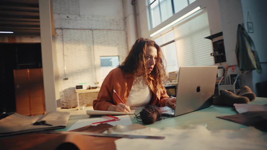 A focused female designer sketches new ideas while researching on her laptop in a sunlit industrial workshop
