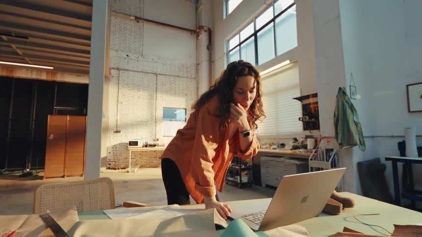 A smiling young female designer works on a laptop and has a video call in her sun-drenched industrial craft studio