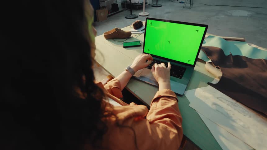 Over-the-shoulder view of a female shoemaker working on a laptop with a green screen in her sunlit artisan workshop