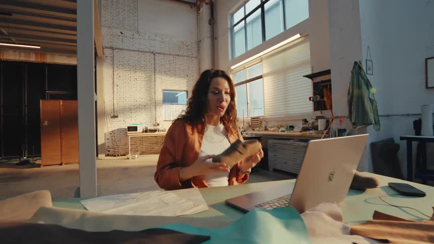 A female footwear designer has a video call on her laptop, showcasing a new shoe design from her sunny workshop