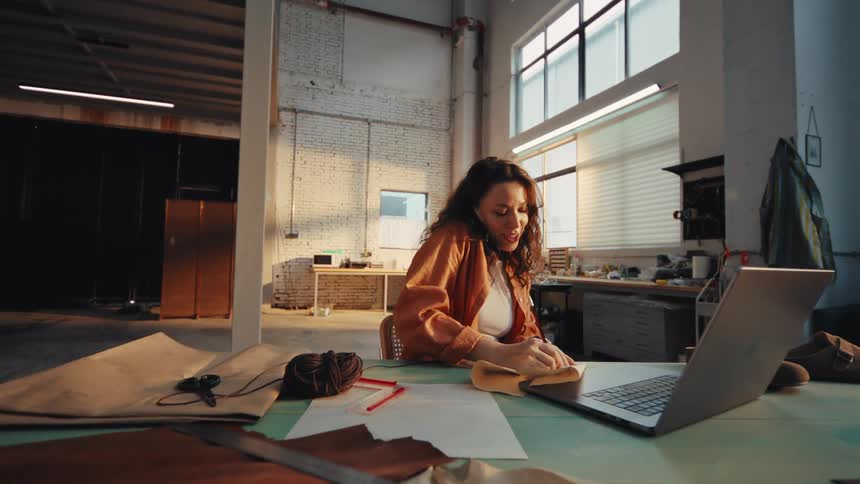 A happy young shoemaker takes a phone call and excitedly begins sketching a new design in her workshop while referencing her laptop