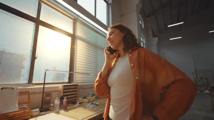 A young female artist stands in her sun-drenched industrial studio, having a thoughtful conversation on her smartphone