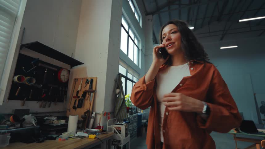 A young female artist with curly hair talks on the phone in her large, sunlit studio