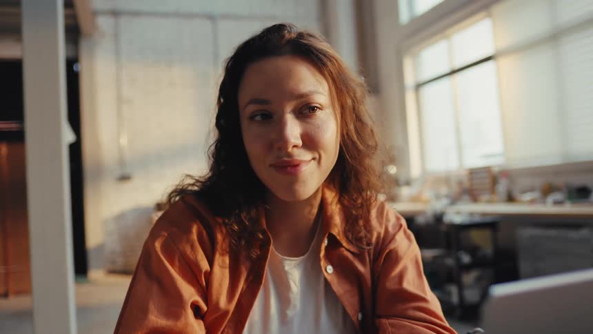 A thoughtful woman with curly hair smiles softly while sitting in her sun-drenched office