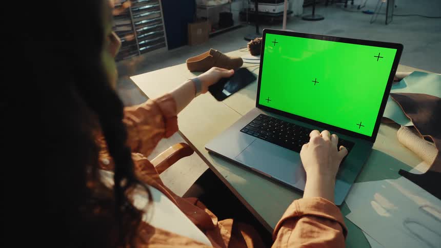 Over-the-shoulder view of a female artisan working on a laptop and holding a smartphone, both with green screens, in her sunlit workshop
