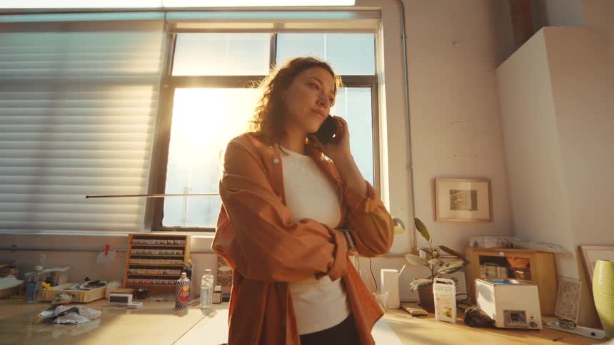 A young artist stands in her sun-drenched studio while listening to a serious phone call