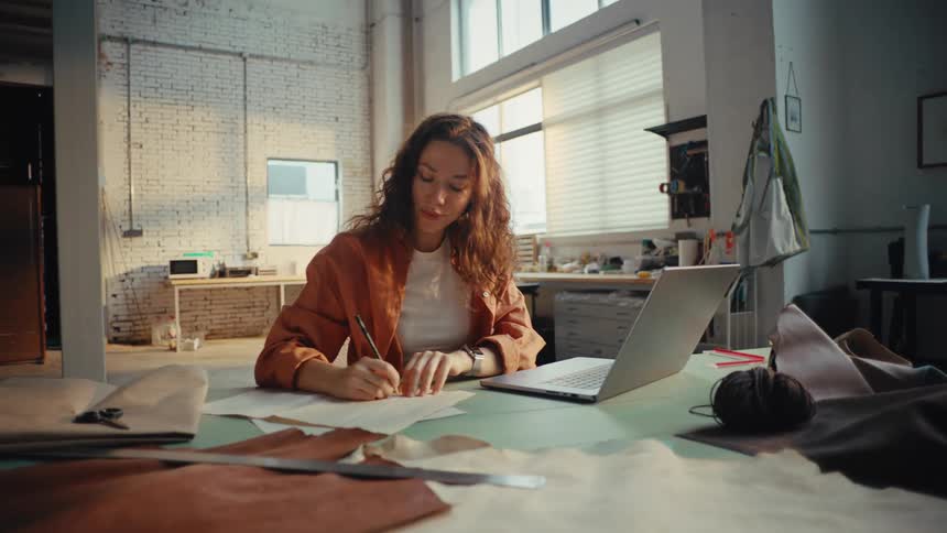 A creative young woman works as a designer in her studio, using a laptop for research and sketching ideas on paper amidst leather samples