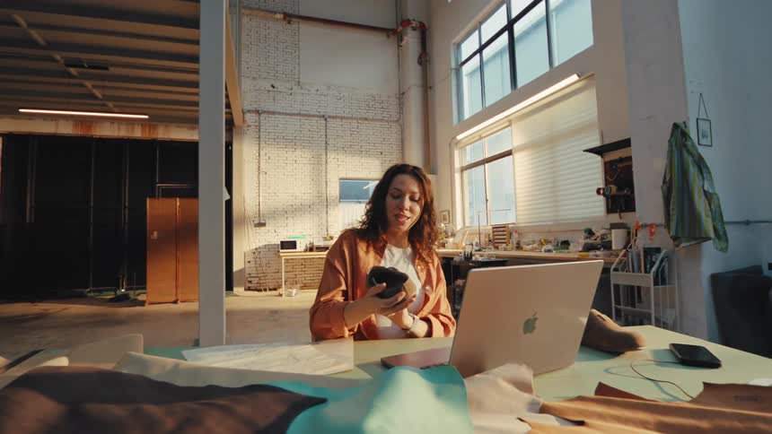 A female shoe designer presents her new handmade footwear design via a video conference call on her laptop from her sunny, spacious workshop