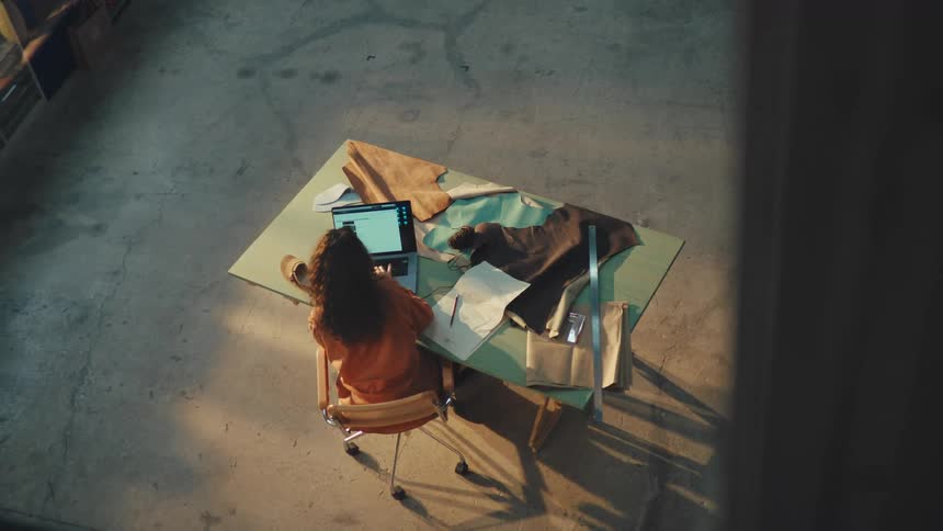 High angle shot of a female designer working on a laptop and sketching a pattern at a workbench with leather and fabric in a studio
