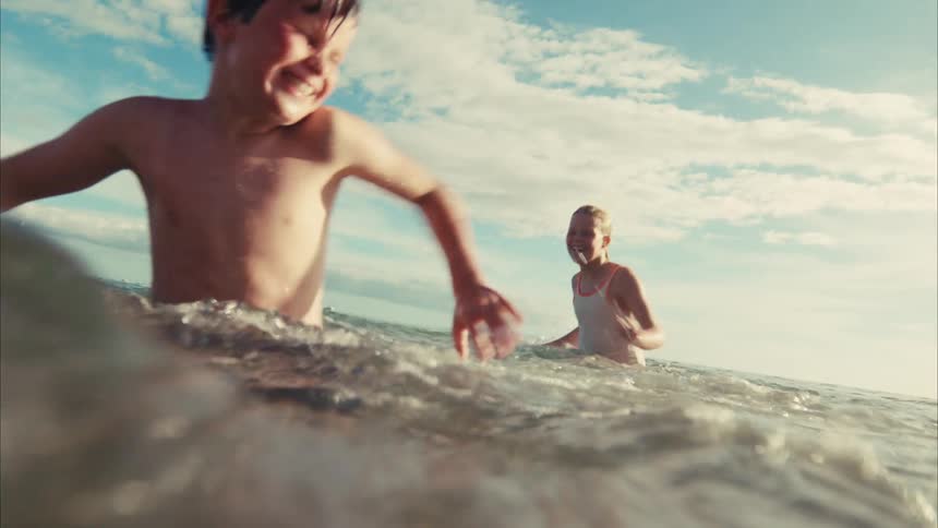 A boy and girl laugh and splash each other playfully in the ocean in a sun-drenched slow-motion shot from the water's surface