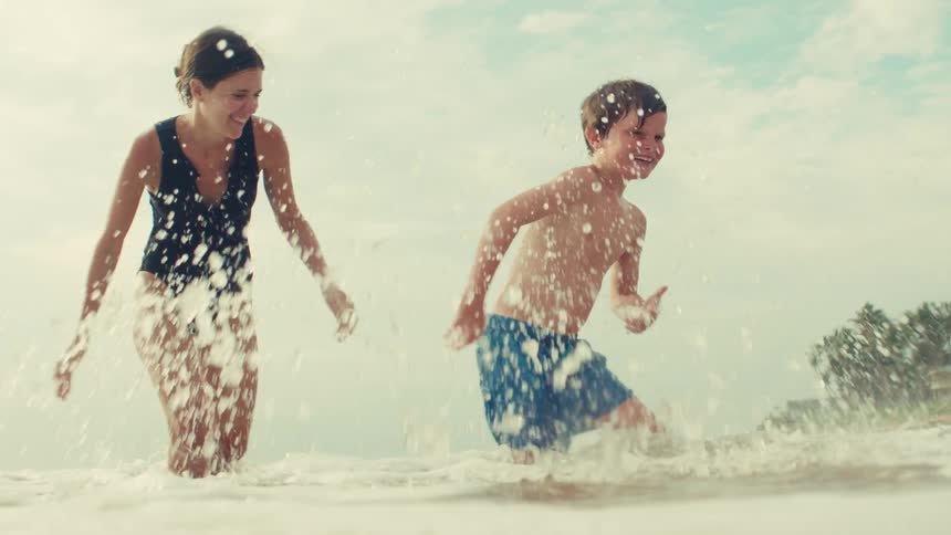 In an epic slow-motion shot, a mother and her son laugh and play as a wave splashes around them on a beautiful beach