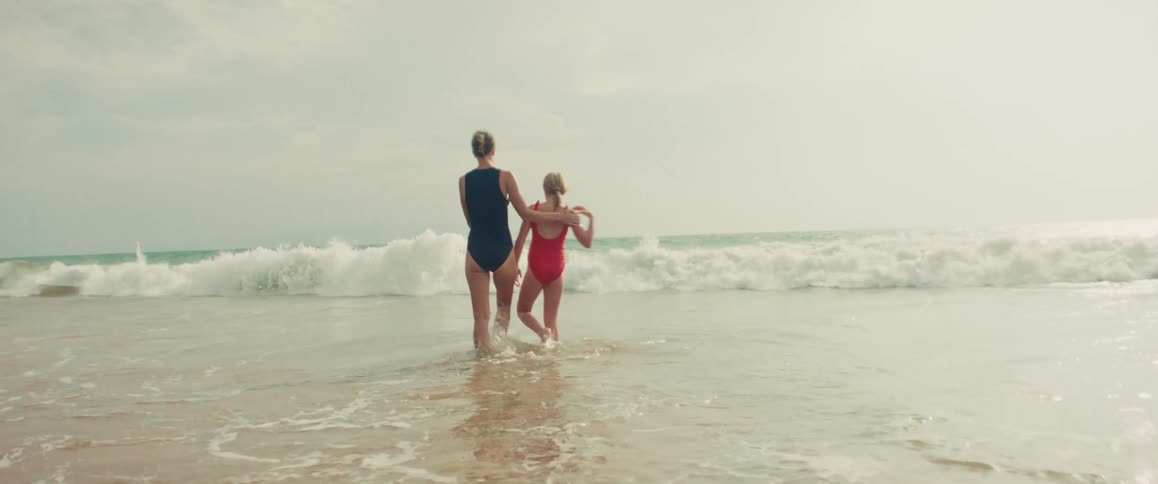 A mother and daughter share a loving embrace while standing in the ocean shallows, watching the waves roll in during a slow motion shot