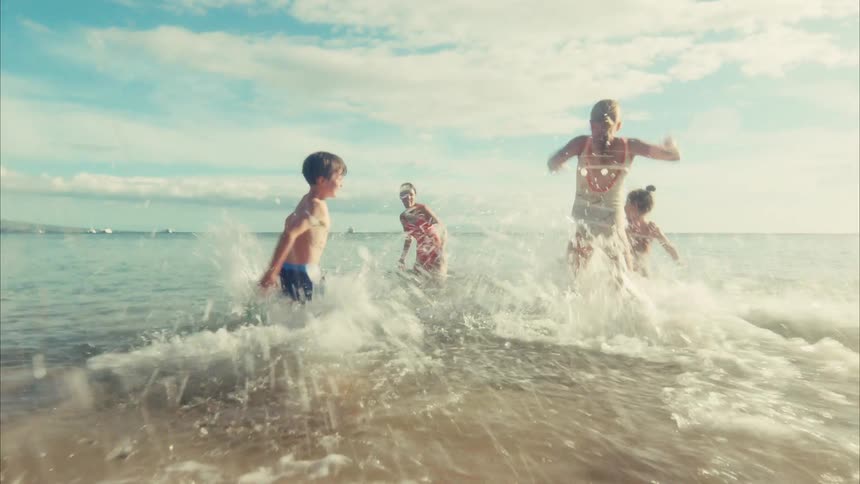 Joyful children run in slow motion across a sunny beach and splash into the shallow ocean waves