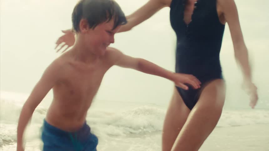 A happy young boy splashes water towards the camera in slow motion while playing with his mother in the ocean waves
