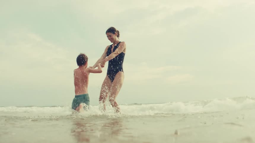 A happy mother and her son playfully spin and splash together in the gentle ocean waves in slow motion