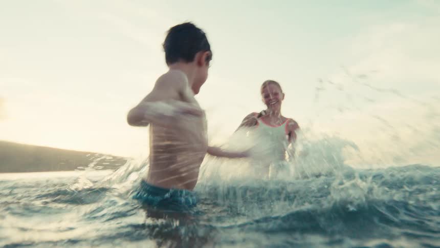 A young boy and girl laugh and joyfully splash each other while playing in the ocean at sunset in slow motion