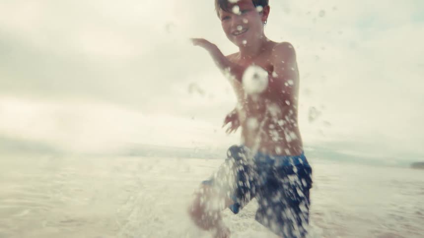 Joyful young boy laughs as he splashes water towards the camera while running in the ocean in slow motion