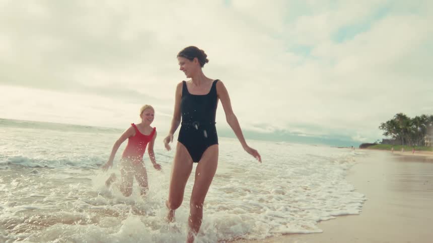 A happy mother and her daughter playfully run through the shallow ocean waves on a sandy beach in slow motion