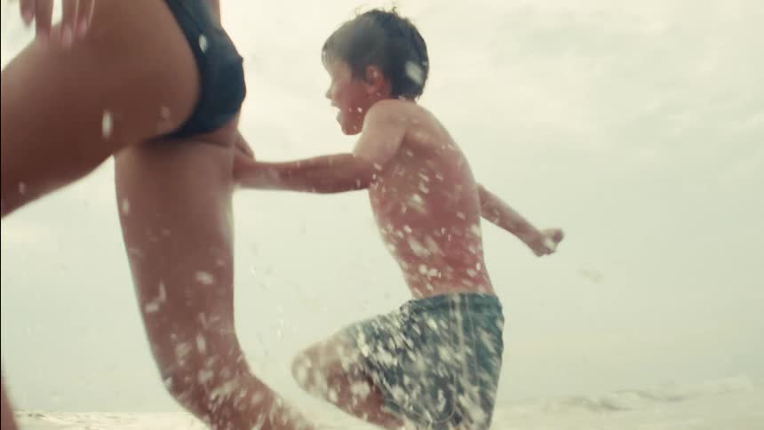 A mother and her young son run hand-in-hand, splashing through the ocean waves in slow motion