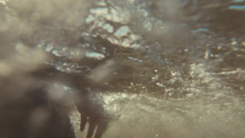 A family plays together in the ocean on a sunny day, with a young boy splashing water in slow motion as the camera goes underwater