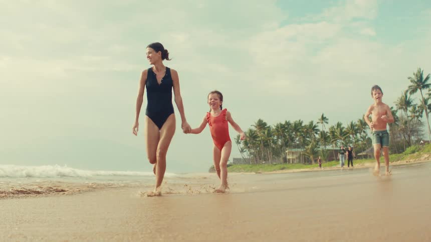 A joyful mother and her two young children hold hands and run along the shoreline of a tropical beach in slow motion