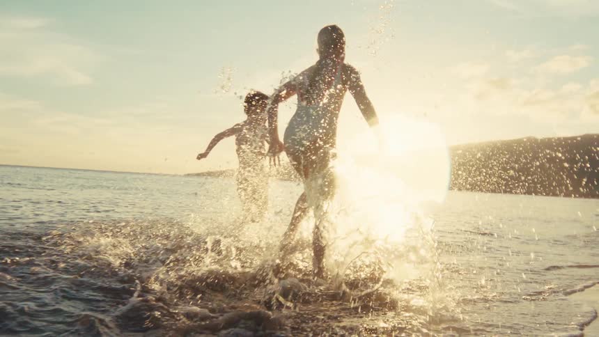 A mother and her young son joyfully run and splash in the shallow ocean water at sunset in beautiful slow motion