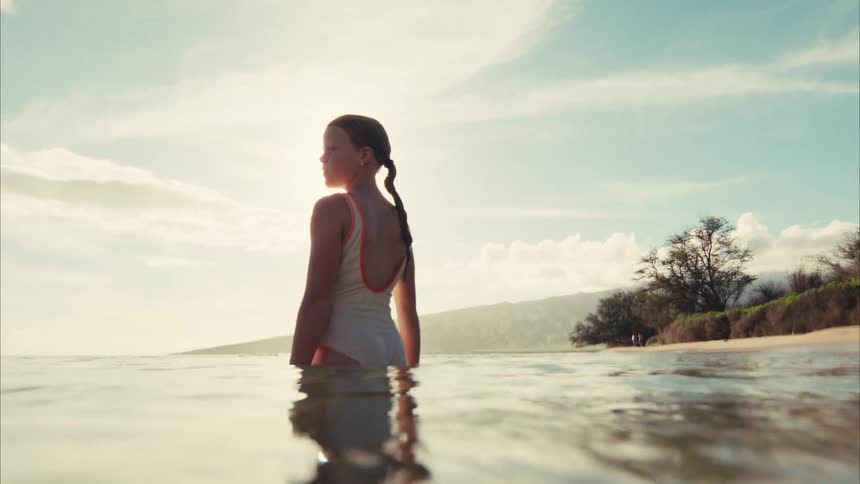 A serene over-under shot shows a young girl in a swimsuit standing peacefully in the calm ocean at a sun-drenched tropical beach