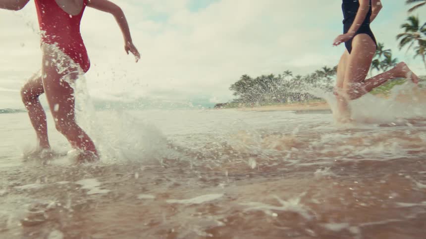 Two joyful young women in swimsuits run and splash through the shallow ocean water on a tropical beach in slow motion
