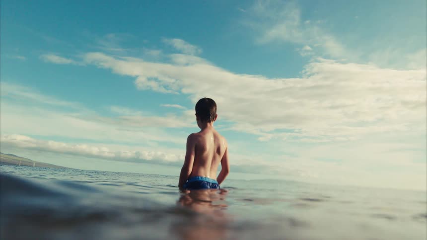 From the water's surface, a young boy contemplatively watches the horizon from the calm ocean in slow motion