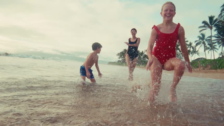 A mother and her two young children run and splash playfully in the shallow ocean waves on a tropical beach in slow motion