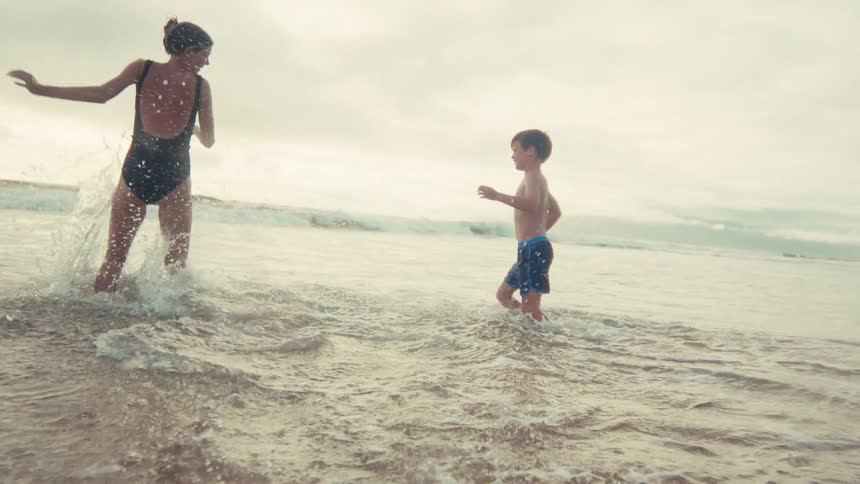 A mother and her young son joyfully run and splash in the shallow ocean waves in super slow motion